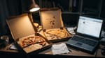 Zwei angebissene Pizzen in offenen Kartons auf einem Schreibtisch neben einem alten Laptop im Lampenlicht