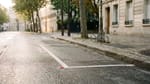 Ein leerer, weiß markierter Parkplatz am Straßenrand einer europäischen Stadt im Morgenlicht, nasser grauer Asphalt, im unscharfen Hintergrund Platanen und Altbaufassaden.