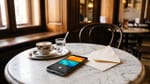 Smartphone auf einem Marmortisch im Wiener Kaffeehaus, auf dem Display zwei bunte Kacheln und eine dunkle mit Schloss, daneben Melange, Wasserglas und ein geöffnetes Kuvert.