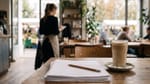 Ein Stapel Papier mit Bleistift und ein Café Olé auf einem Holztisch, im Hintergrund eine Kellnerin mit Bestellblock vor dem Markt.