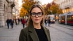 Porträt einer jungen Frau mit schwarzer Datenbrille auf einer Wiener Herbststraße, im rechten Glas eine kleine Anzeige, im Hintergrund fährt eine Straßenbahn.