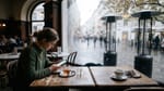 Junge Frau mit Handy an einem Kaffeehaustisch, daneben ein leerer Tisch, draußen ausgeschaltete Heizstrahler im Oktoberregen
