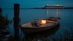 Ein unbeschriftetes Ruderboot liegt in der Dämmerung am Ufer vertäut, auf der Bank leuchtet ein aufgeklappter Laptop; weit draußen zieht ein hell erleuchtetes Kreuzfahrtschiff vorbei.