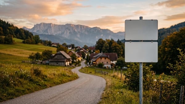Zwei gleich große, leere weiße Tafeln an einem Pfosten am Ortseingang, dahinter eine Landstraße, ein Dorf und Berge im Abendlicht.
